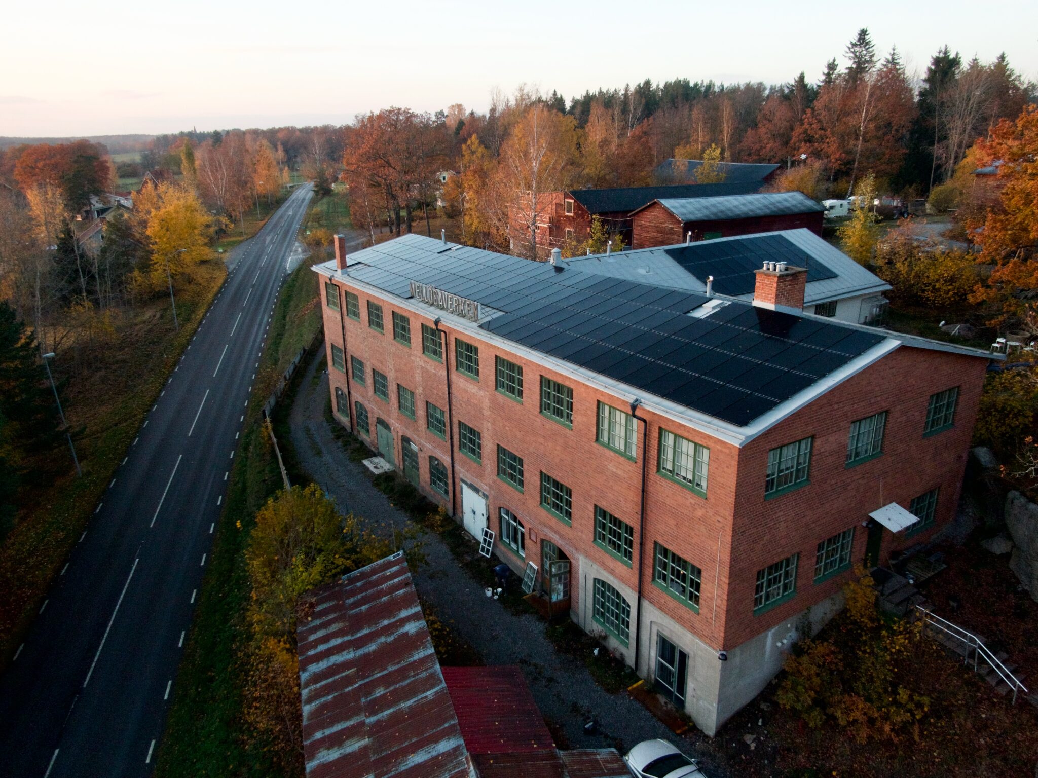 Aerial view of a brick building with solar panels, surrounded by autumn trees and a road.