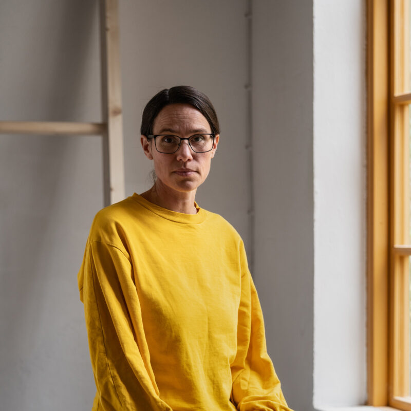 Woman in a yellow sweatshirt and glasses sitting against a white wall with a wooden shelf and window.