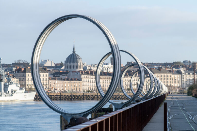 Metal rings by Daniel Buren along the Quai des Antilles, with a cityscape and dome in the background.