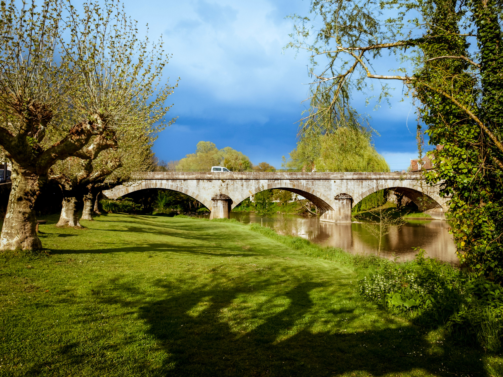 Stone bridge with arches over a river, surrounded by trees and grassy area under a cloudy sky.