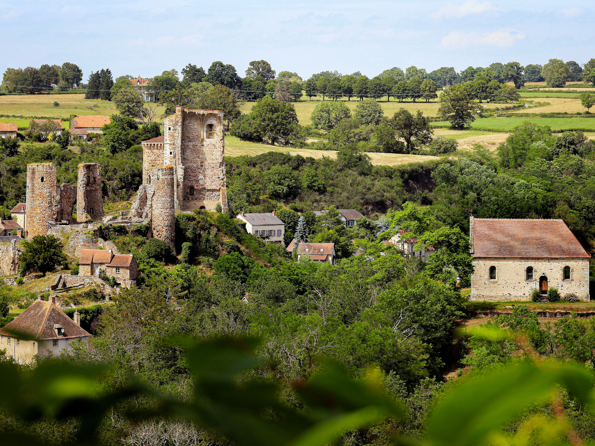Ruins of a medieval castle surrounded by lush greenery and small houses, under a blue sky with scattered clouds.