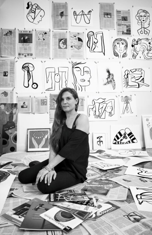 Hanna Barczyk sits on the floor surrounded by her artwork and newspaper clippings in a vibrant studio.