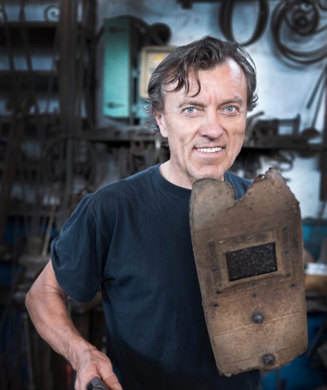 Man holding a metalworking tool and a protective shield, smiling in a workshop filled with tools and equipment.
