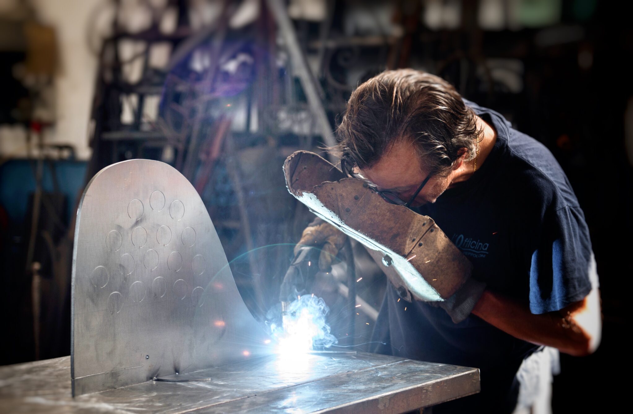 A man in a workshop welds metal, focusing intently as sparks fly from the welding process.