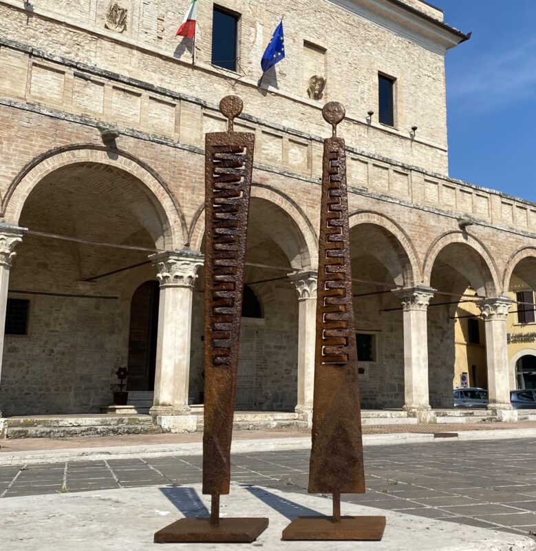 Two tall, rust-colored sculptures stand in front of an arched building with flags, under a clear blue sky.