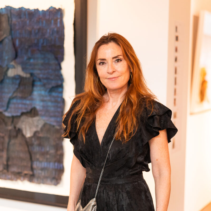 Woman with long brown hair in a black dress stands in an art gallery, with abstract blue artwork in the background.