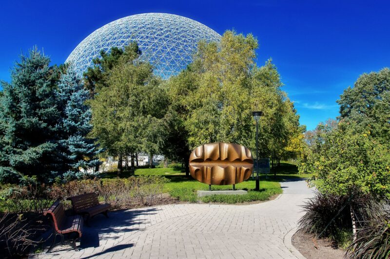 Sculpture in Jean Drapeau Park, Montreal, with trees and the Biosphere dome in the background under a clear blue sky.
