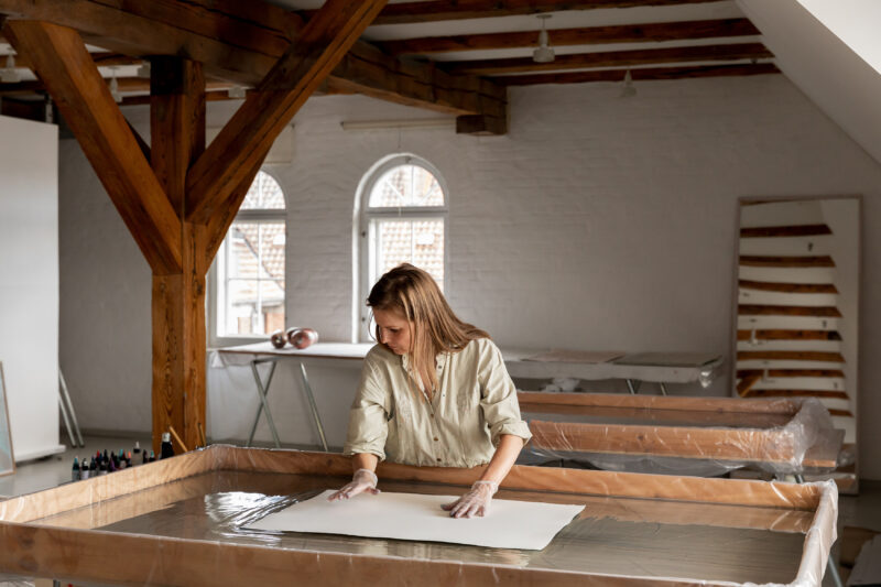 Artist Pernille Snedker Hansen working in a studio, preparing paper on a large table with wooden beams in the background.