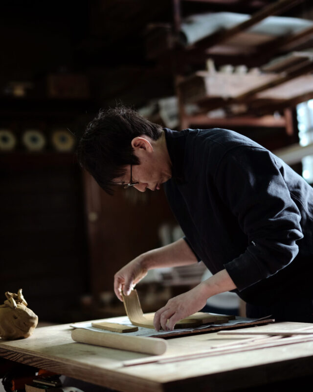 A person in a dark apron carefully rolls dough on a wooden table in a dimly lit workshop.