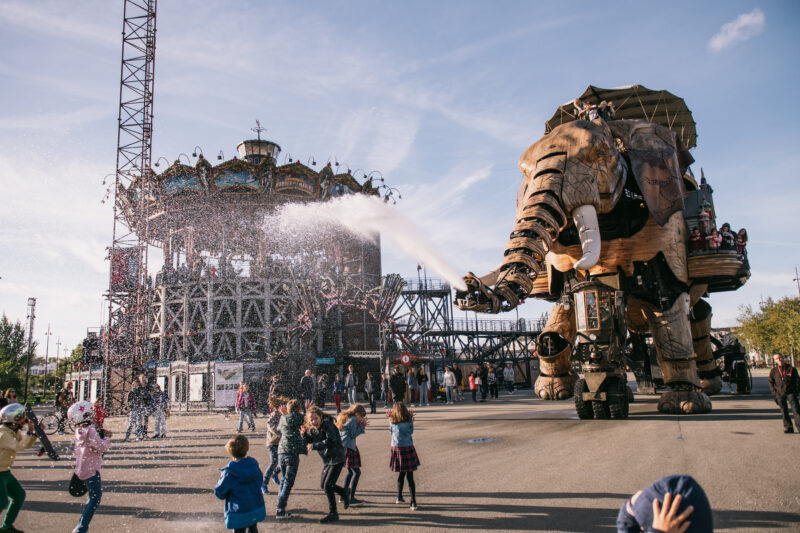 Giant mechanical elephant spraying water in front of a carousel, with children playing nearby at Les Machines de l'île, N...