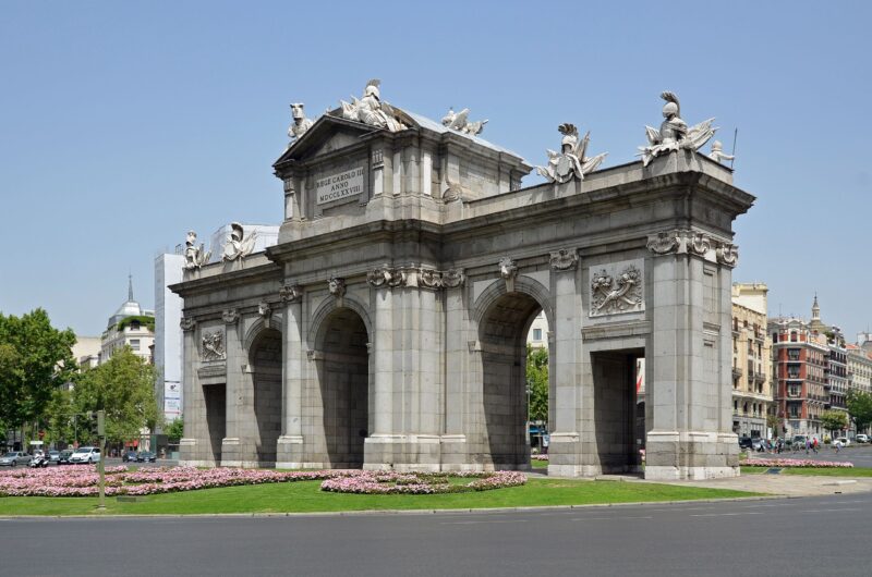 Puerta de Alcalá in Madrid, a large stone archway surrounded by greenery and flowers, under a clear blue sky.