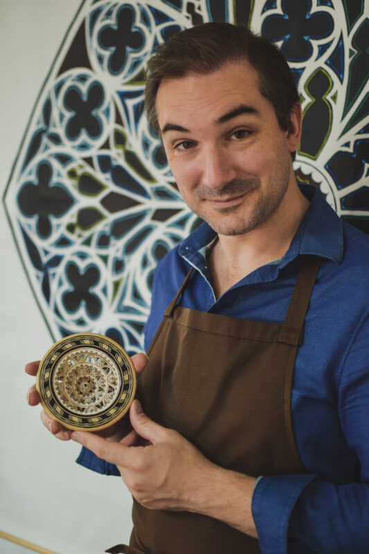Man in a blue shirt and brown apron holds a decorative circular object, smiling in front of a colorful patterned backdrop.