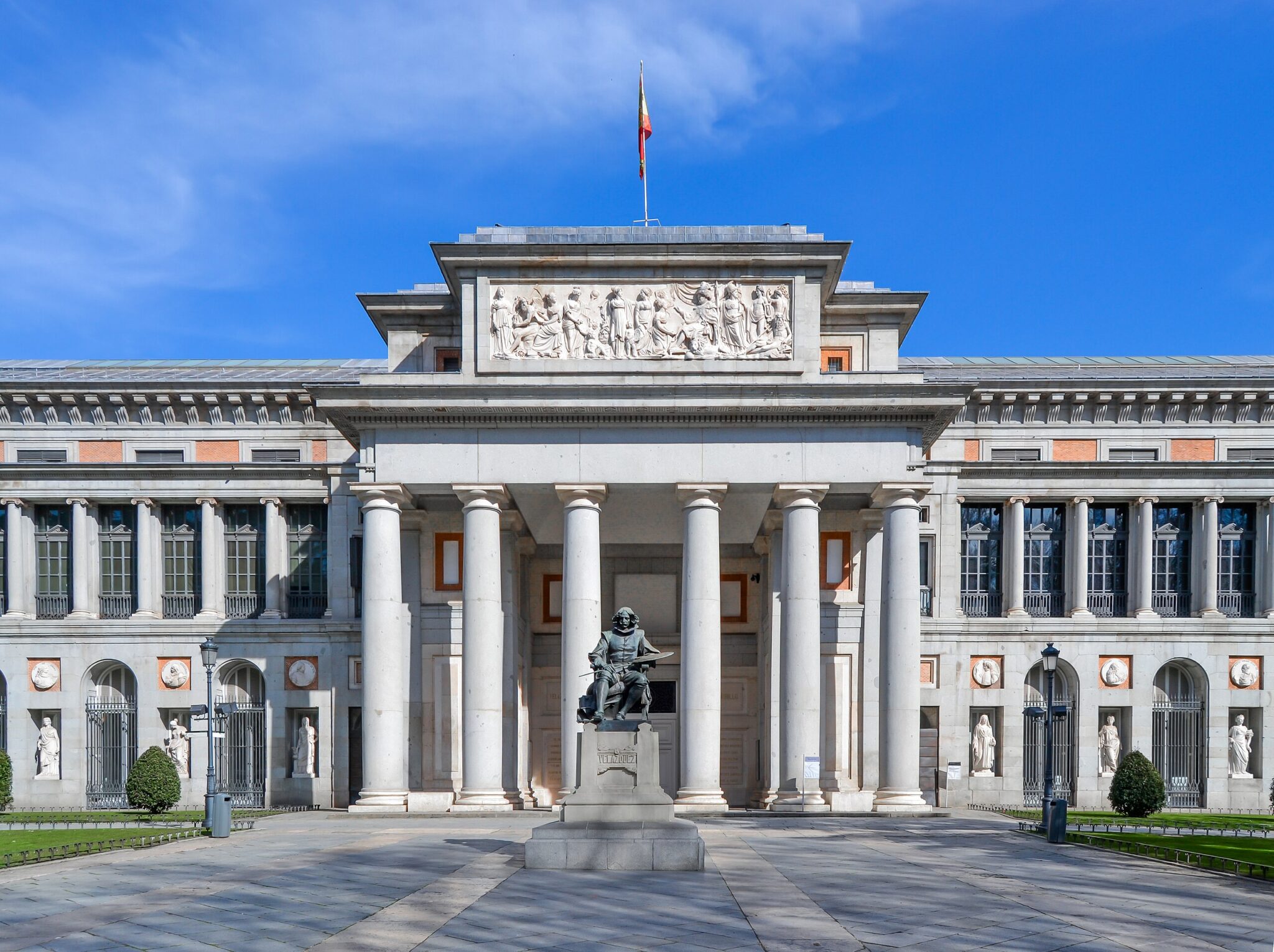 Museo del Prado's grand facade featuring classical columns, sculptures, and a statue in front under a clear blue sky.