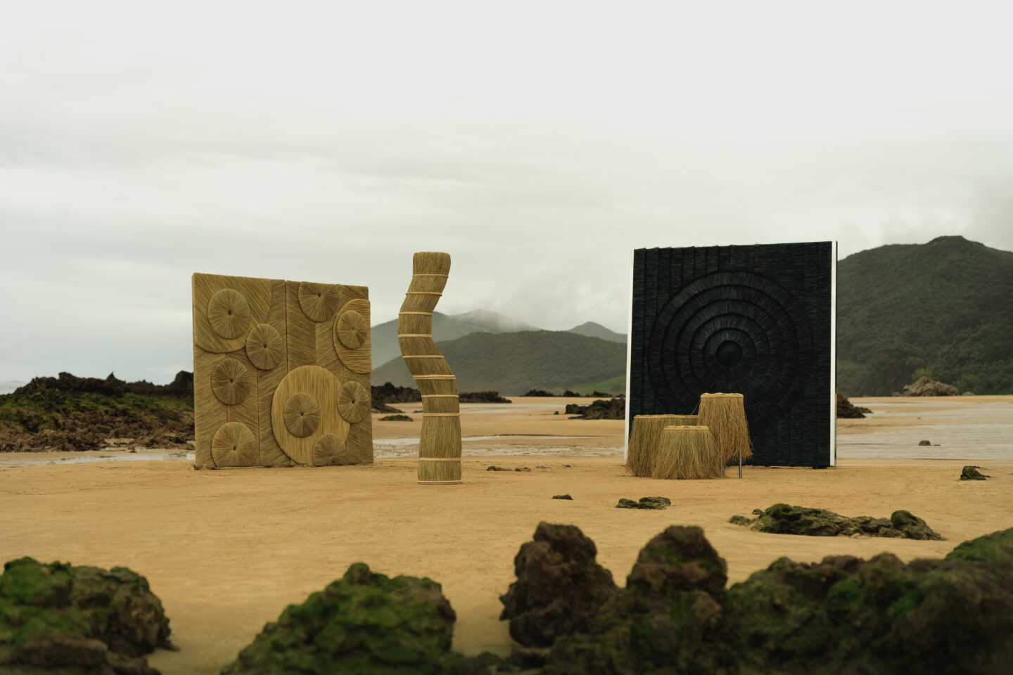 Three abstract sculptures made of natural materials on a sandy beach, with mountains and overcast sky in the background.
