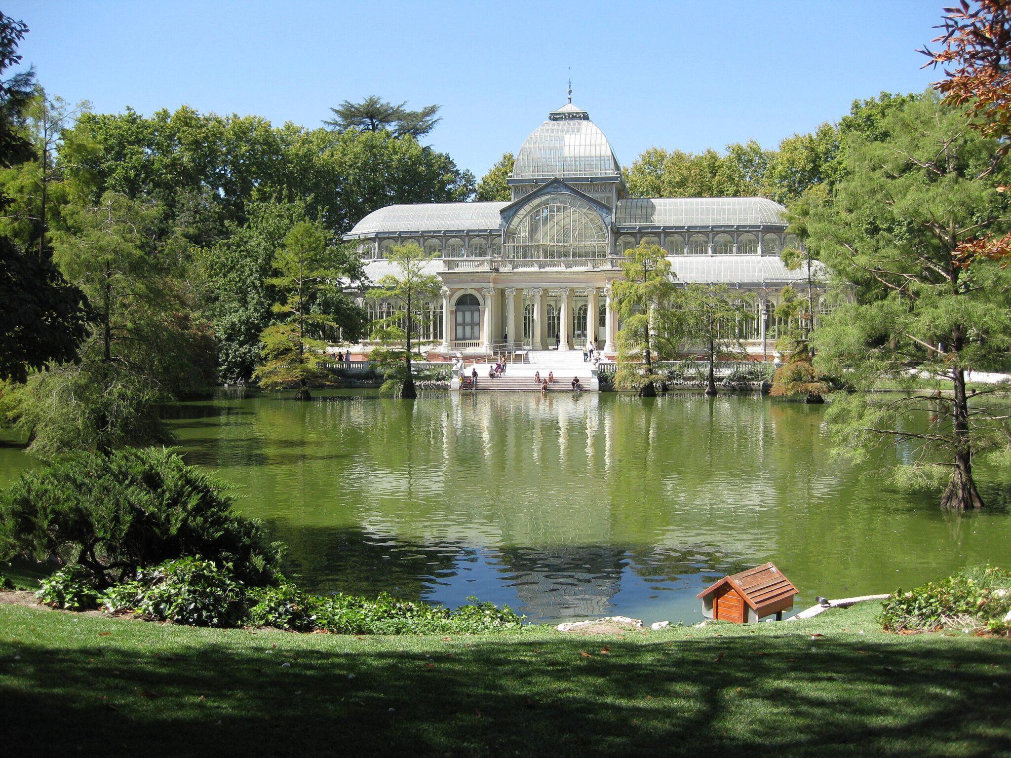 Palacio de Cristal surrounded by greenery, reflecting in a tranquil pond at El Retiro Park, Madrid.