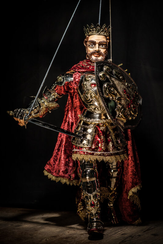 King puppet dressed in ornate armor and a red cape, holding a sword and shield, set against a dark background.