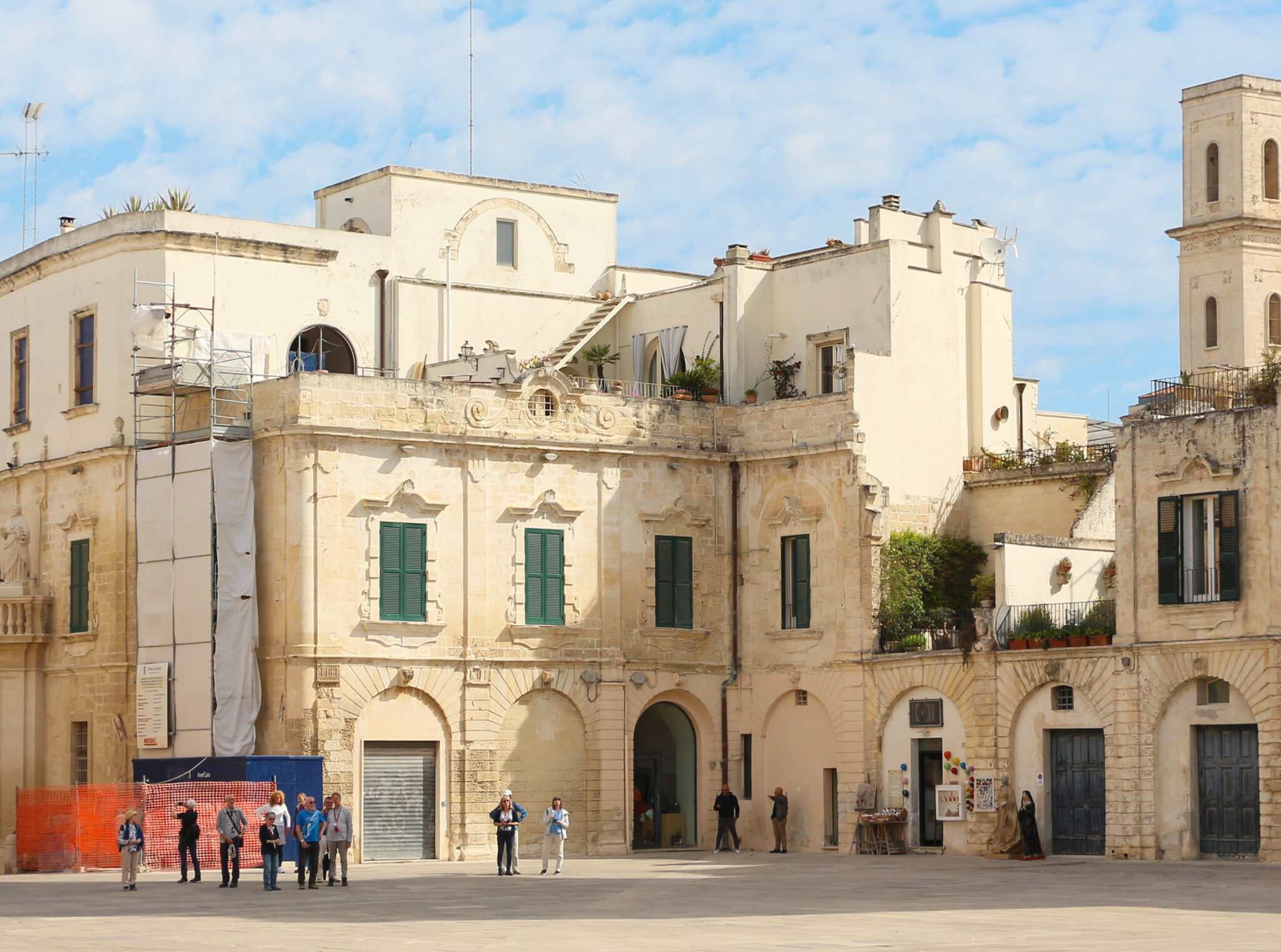 Baroque buildings in Piazza Duomo, Syracuse, with people gathered in the square under a partly cloudy sky.