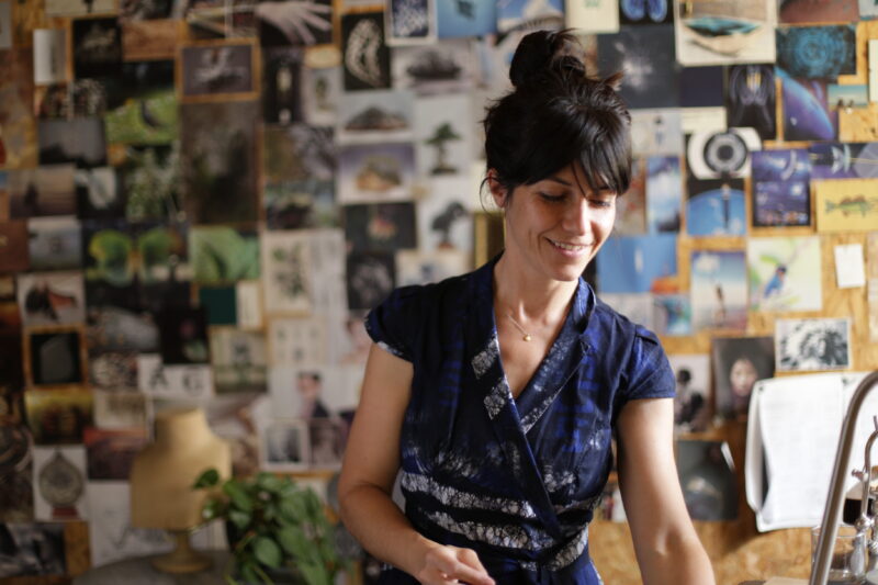 Woman in a blue dress smiles while standing in an art studio filled with photographs and artwork on the walls.