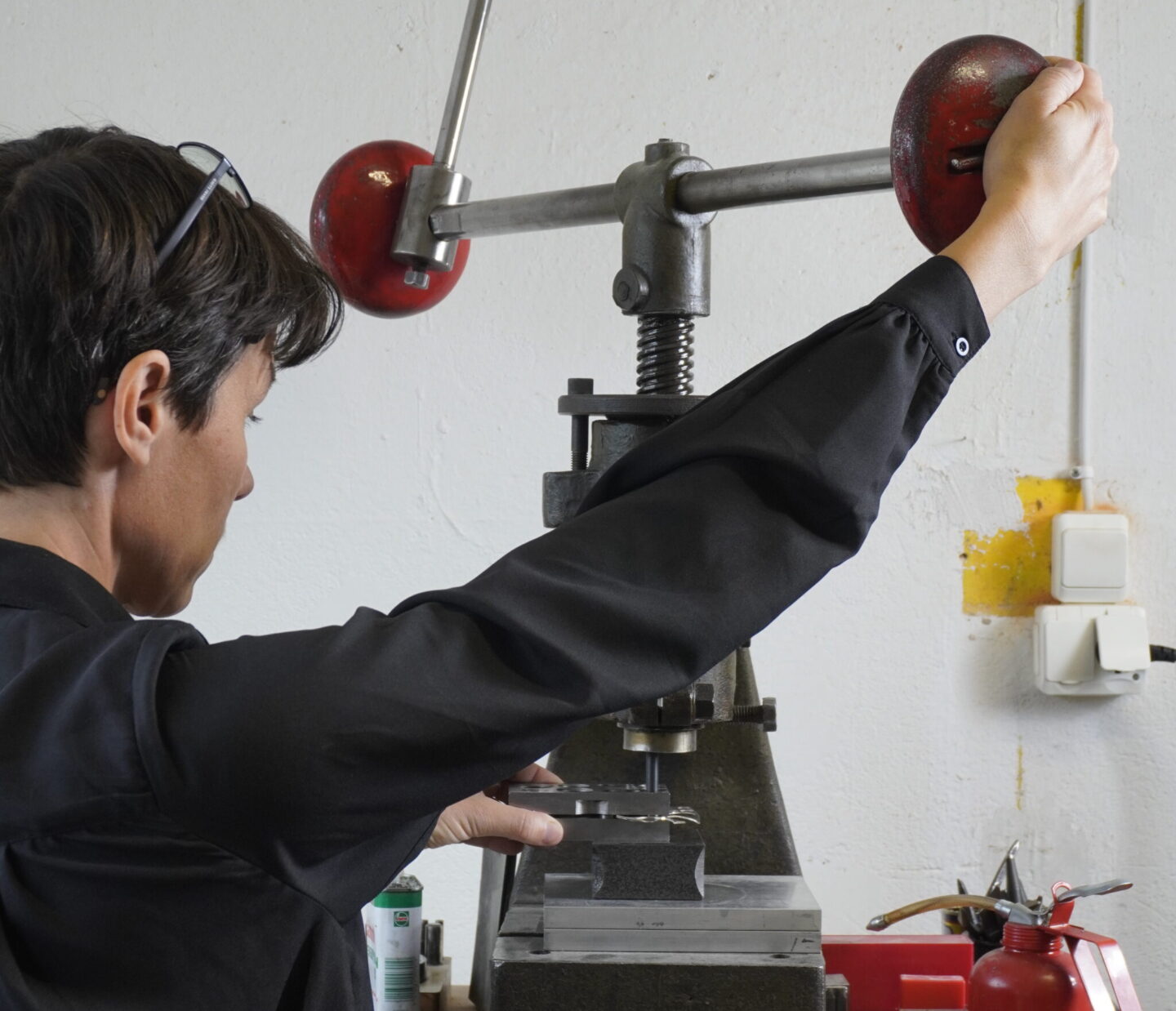 Person operating a metal press machine in a workshop, wearing a black shirt and sunglasses, with tools and equipment nearby.