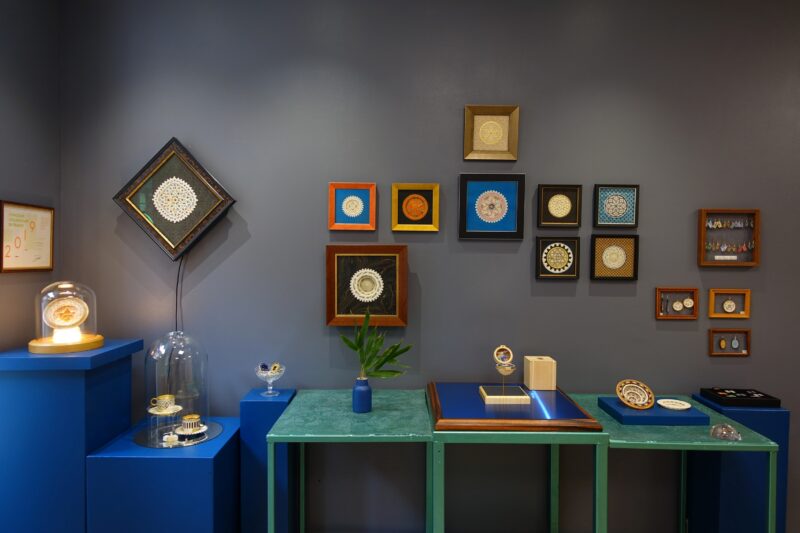 Colorful framed artworks and decorative pieces displayed on a gray wall, with blue tables and a plant in the showroom.