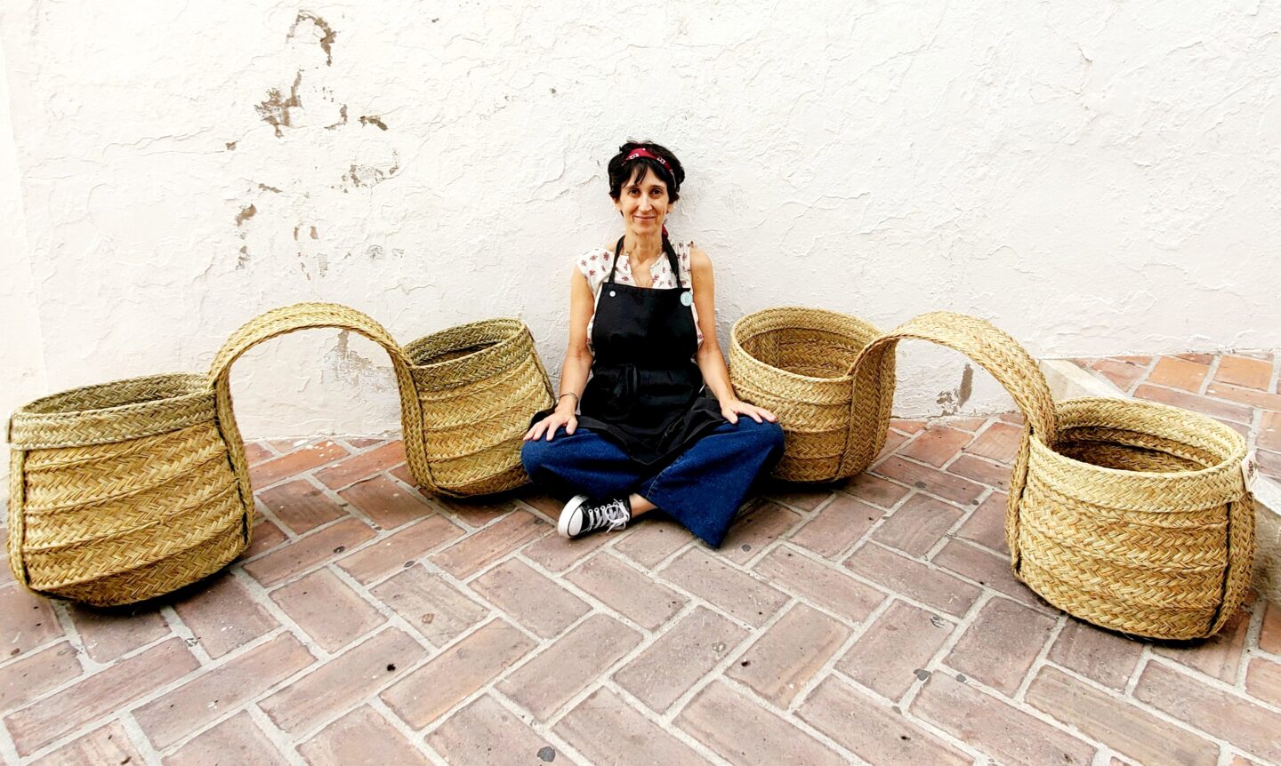 Sonia Lekuona sits on a tiled floor, surrounded by woven baskets, smiling and wearing an apron.