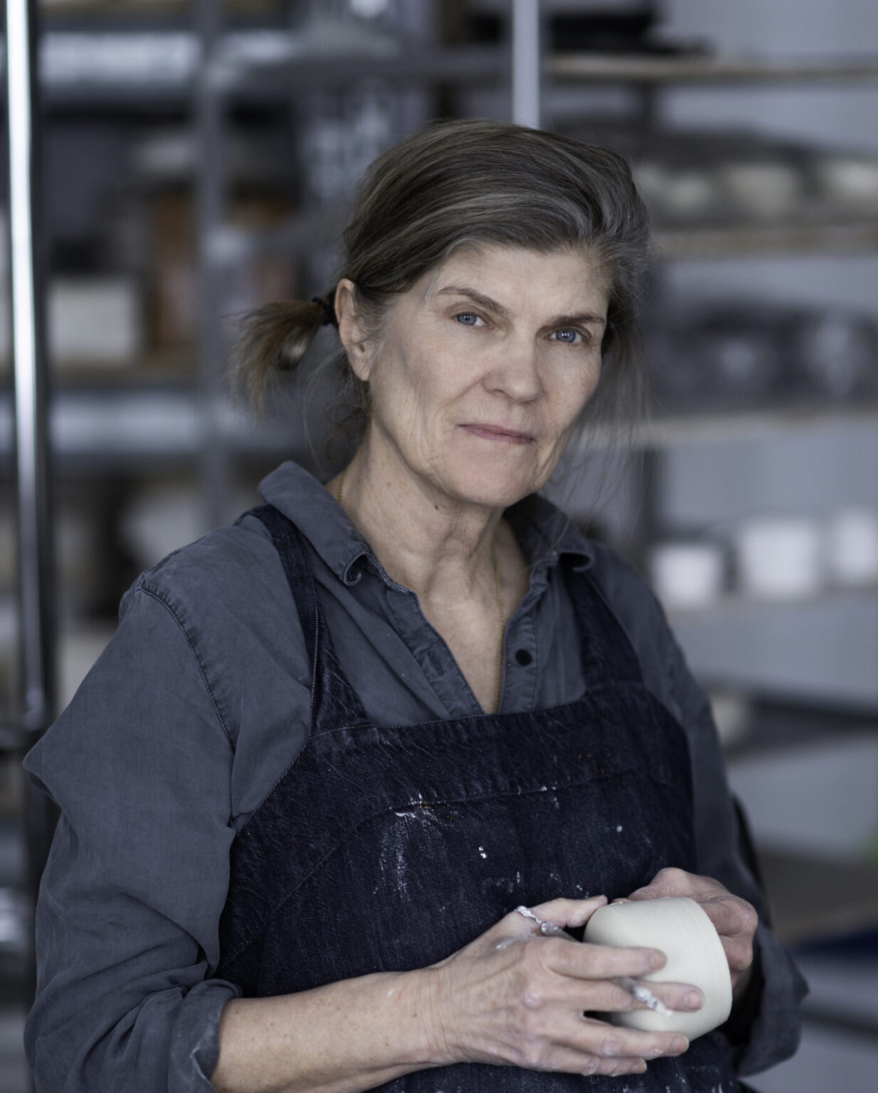Woman with gray hair in a ponytail, wearing a dark shirt and apron, holding a small ceramic piece in a pottery studio.