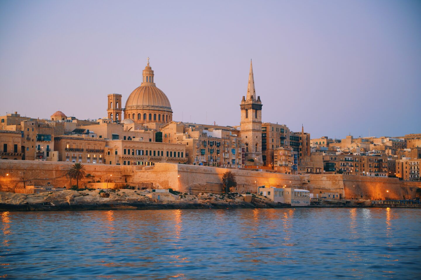 Valletta, Malta at sunset, showcasing the skyline with domed buildings and a spire, reflecting on the sea.