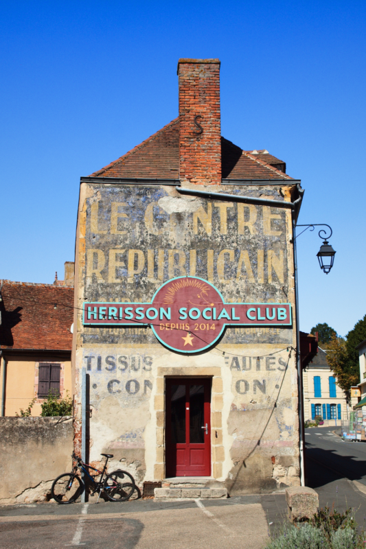Weathered building with a faded sign reading "Le Centre Républicain" and a colorful "Hérisson Social Club" sign, bike pa...