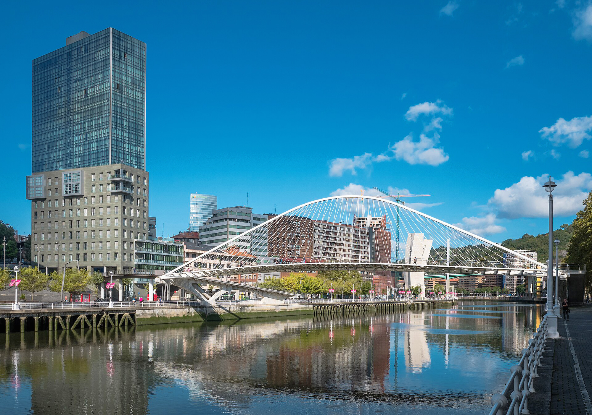 Modern Zubizuri bridge spans the Nervión River, with urban buildings and a clear blue sky in the background.