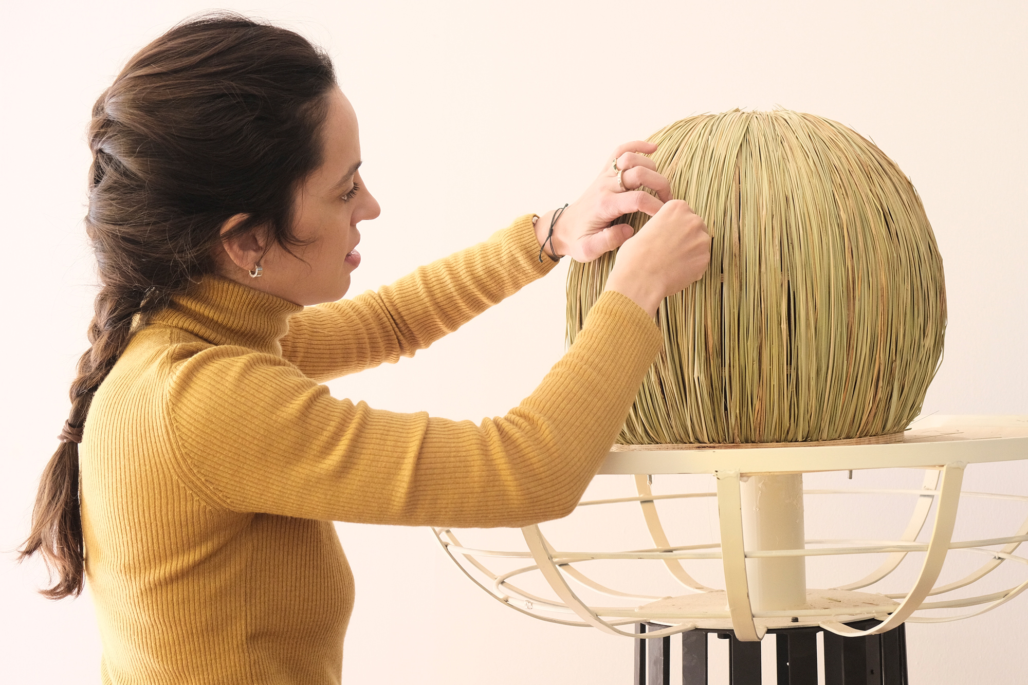 Woman in a mustard turtleneck assembling a round straw sculpture on a stand.