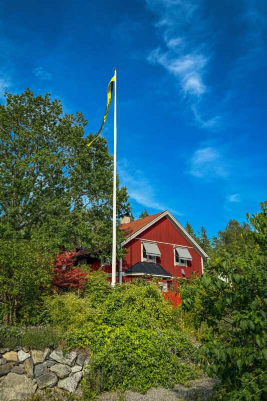Red house with a yellow flag on a pole, surrounded by greenery and trees under a blue sky.
