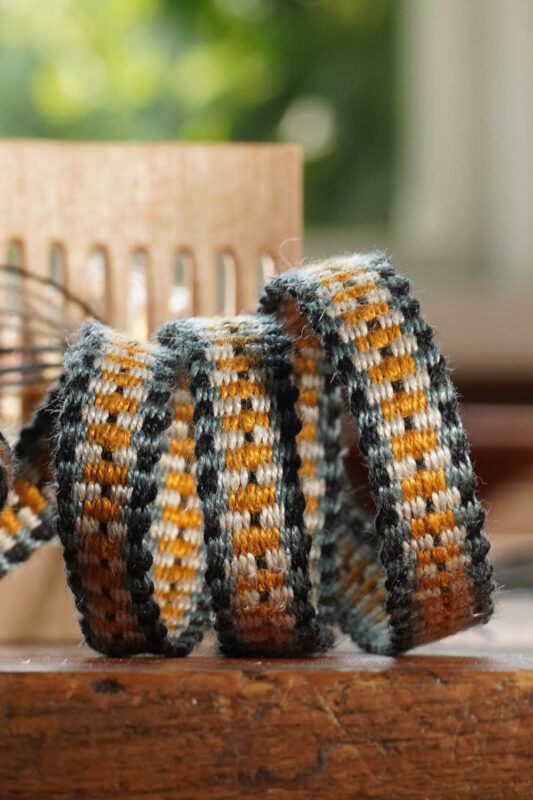 Woven ribbon in orange, black, and gray patterns, coiled on a wooden surface with a blurred background.