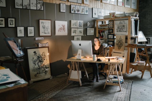 A woman works at a desk in an art studio surrounded by framed artworks and sketches on the walls.