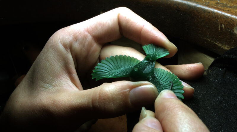 A person holds a green wax bird sculpture, carefully detailing its features with a small tool.