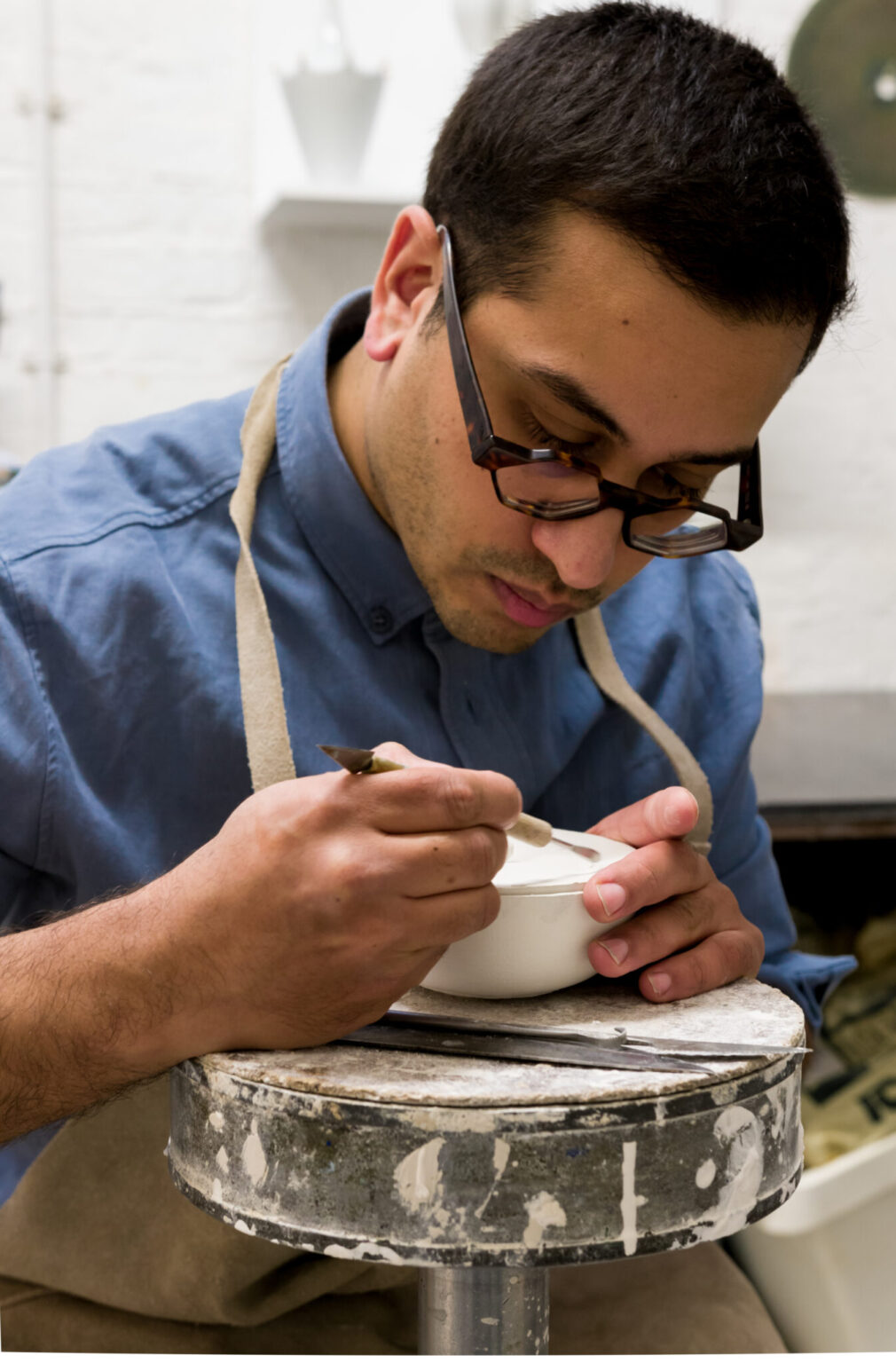 A man with glasses carefully paints a small bowl while seated at a workbench, wearing an apron.