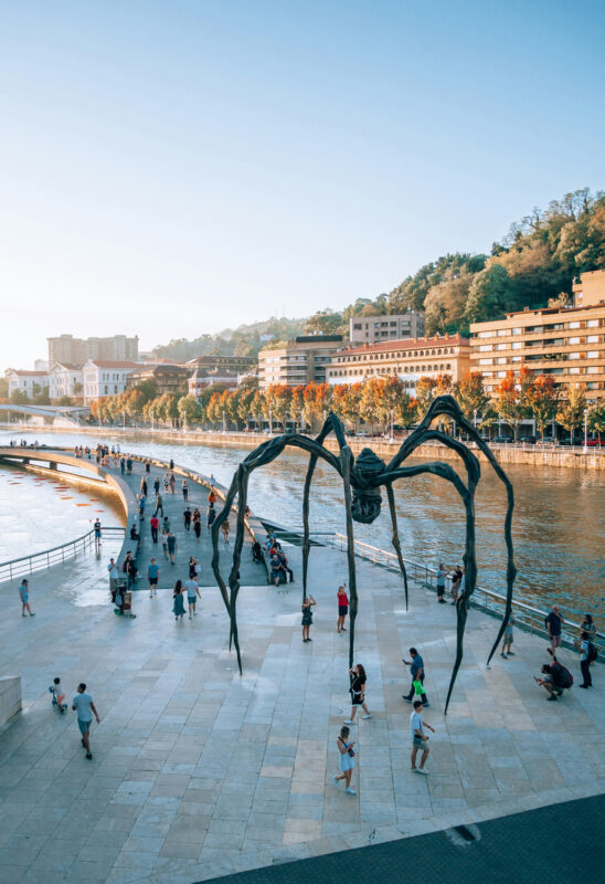 Giant spider sculpture on a riverside promenade with people walking and buildings in the background.