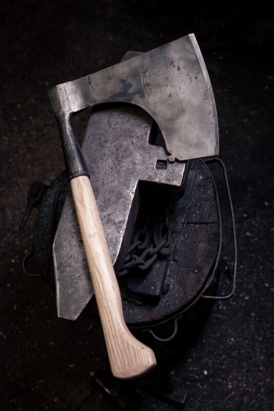 A metal axe with a wooden handle rests on an anvil, surrounded by tools and chains, in a workshop setting.