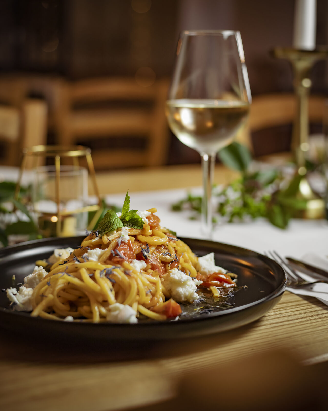Plate of pasta topped with tomatoes and herbs, accompanied by a glass of white wine and a candle on a dining table.
