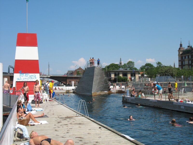 Islands Brygge Harbour Bath with swimmers, wooden platforms, and a red and white striped tower under a clear blue sky.