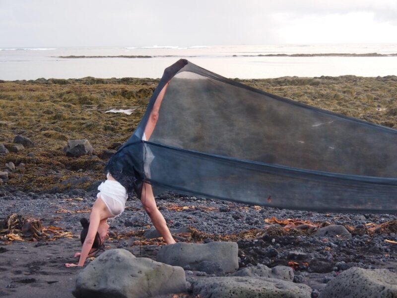 A dancer in a flowing outfit performs a handstand on a rocky beach, with ocean waves in the background.