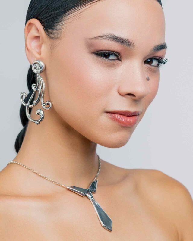 Model showcasing handcrafted silver jewelry, featuring an ornate earring and a geometric necklace, with a neutral background.