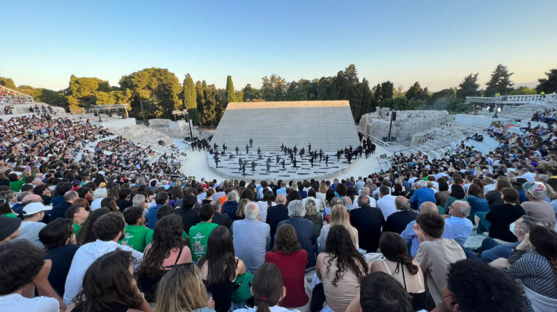 Performance at the Greek Theatre of Syracuse, with a large audience watching on a sunny day.