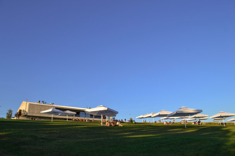 Lawn with people relaxing under white umbrellas, overlooking the Kabinet Brewery building against a clear blue sky.