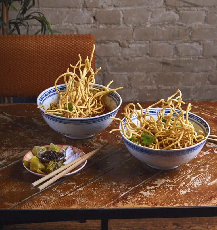Two bowls of Khao Soi with crispy noodles, garnished with cilantro, on a wooden table with chopsticks and condiments.