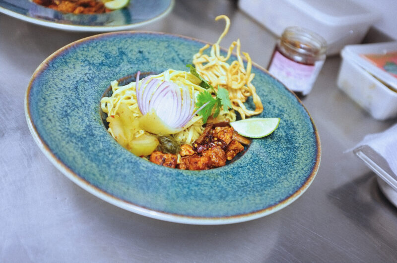 Bowl of Khao Soi featuring yellow noodles, tofu, lime, and garnished with onion and herbs on a blue plate.