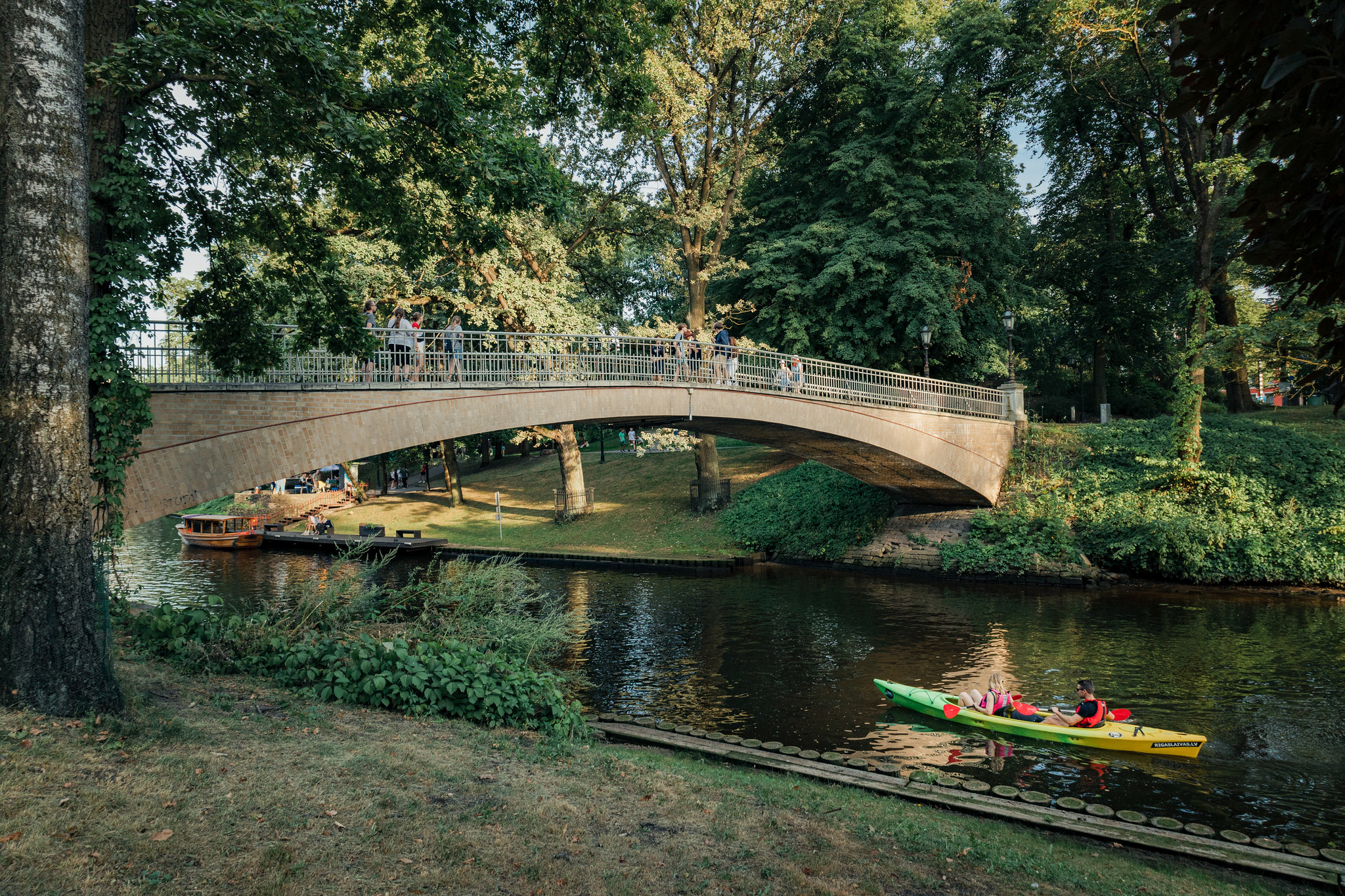Bastejkalns Park in Riga with a bridge, people walking, and a kayak on the calm water.