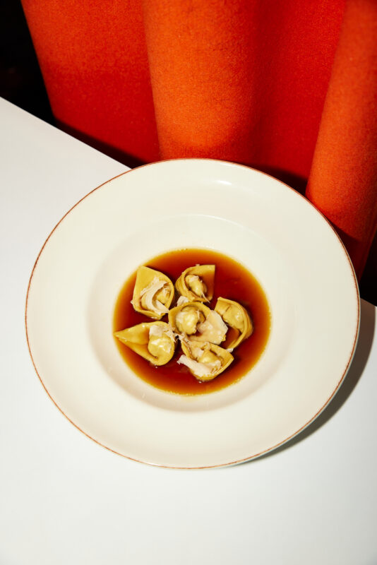 Dumplings in broth served in a white bowl on a table, with an orange curtain in the background.