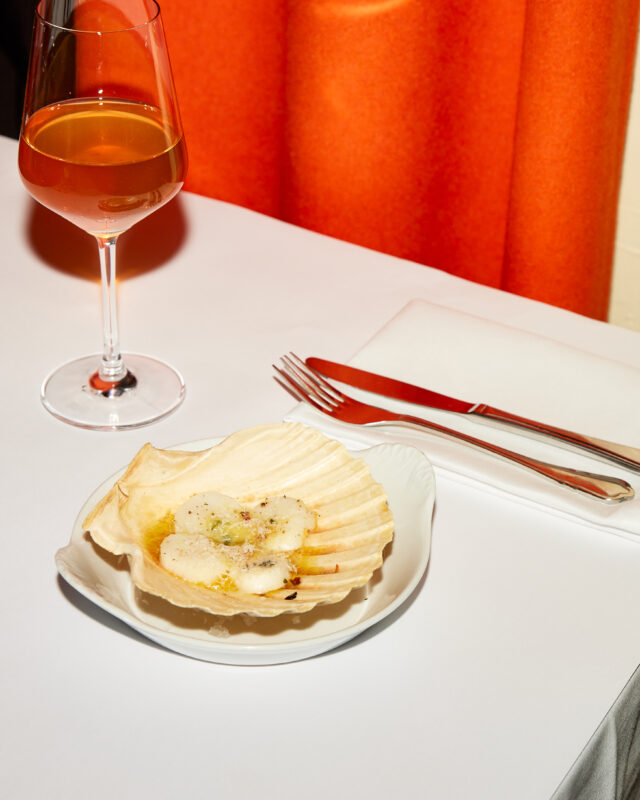 A scallop dish on a white plate beside a glass of orange wine, set against an orange curtain backdrop.