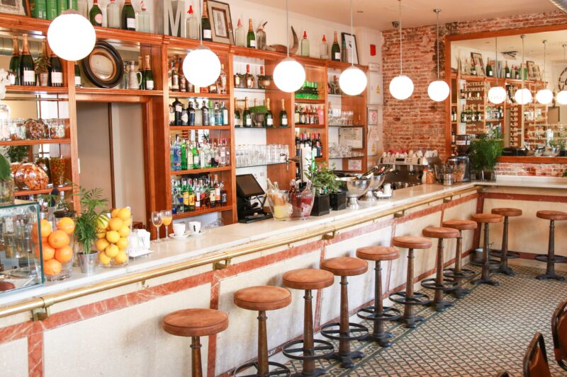 Bar counter at Murillo Café with wooden stools, shelves of drinks, and decorative lighting.