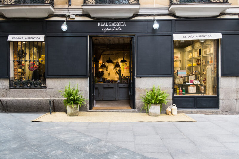 Entrance of Real Fábrica Española store, featuring potted plants and displays in windows.